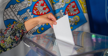 Zdjęcie przedstawia rękę wrzucającą kartę do głosowania do urny wyborczej. W tle flagi z herbem Miasta Poznania.