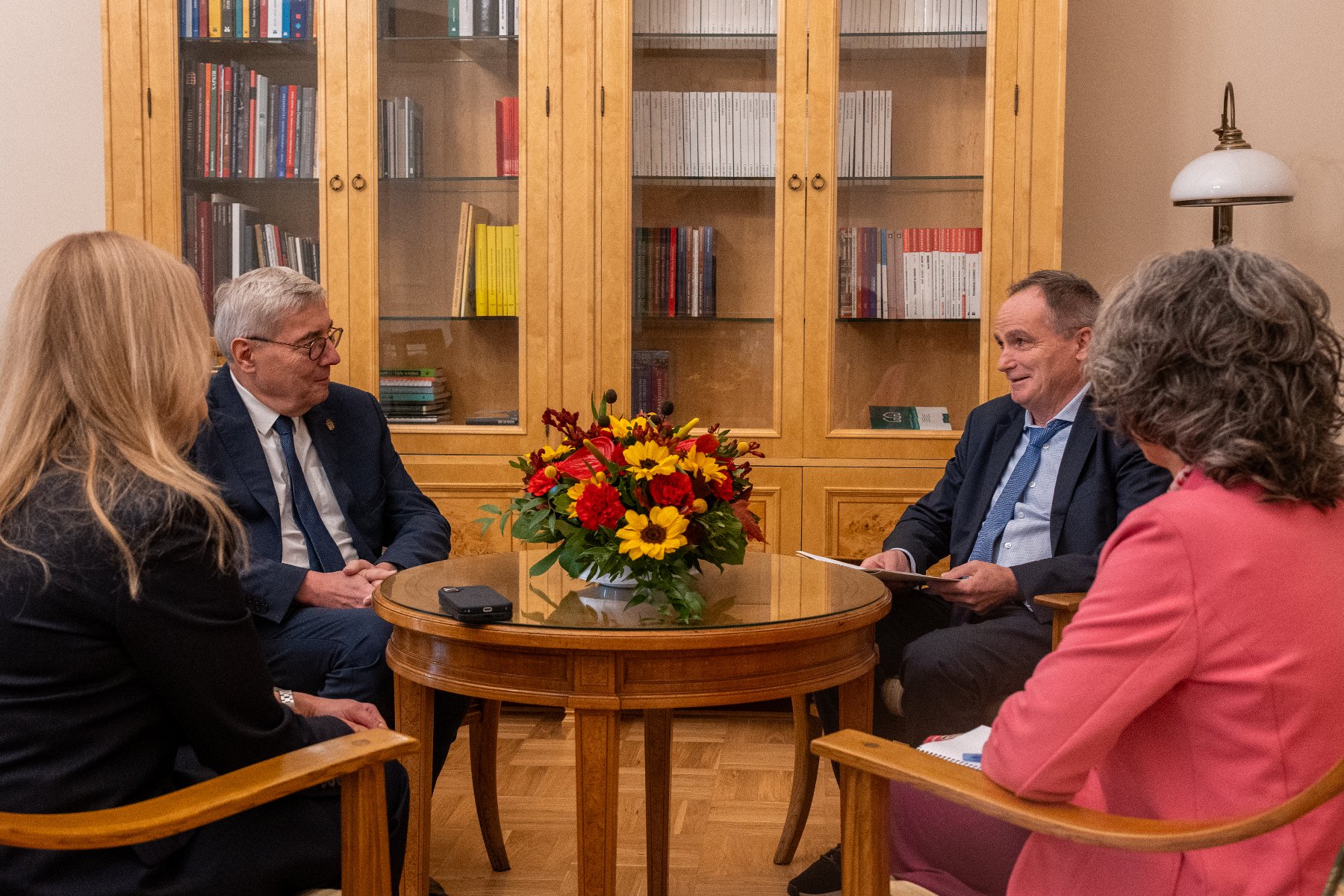 Spotkanie czterech osób przy okrągłym stole z bukietem kwiatów w gabinecie z drewnianą biblioteczką w tle.
